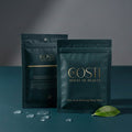 Two Dr. Costi sheet masks on a dark surface with a leaf and water droplets.